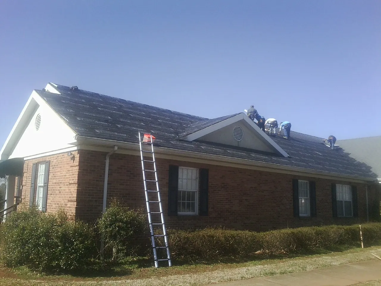 Crew tearing shingles off a brick ranch roof with a ladder set — Metal Roof Repair work by our crew in Clark