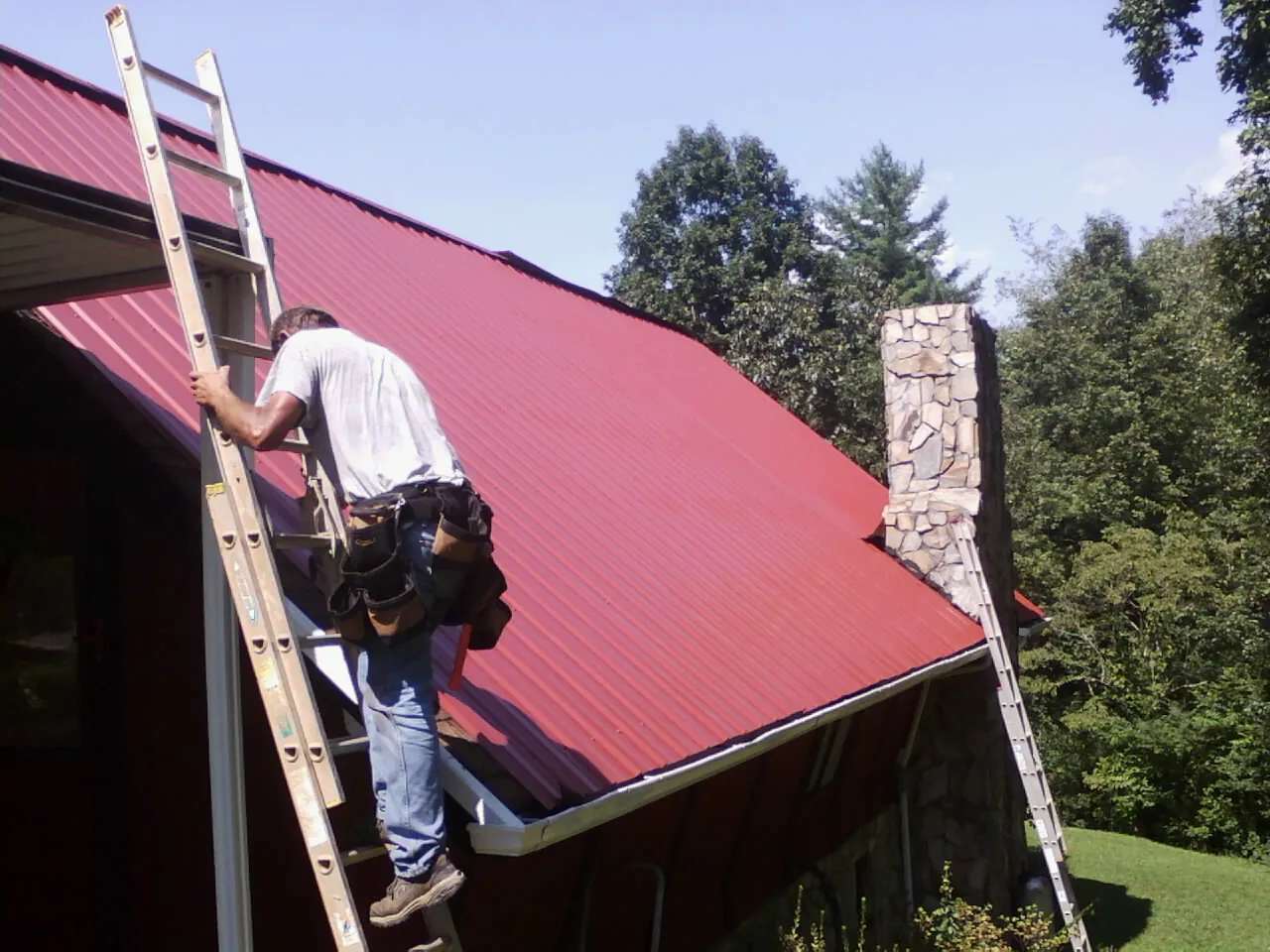 Roofer on a ladder beside a red metal roof and stone chimney — Soffit & Fascia Repair by Roofing Brothers in Clark